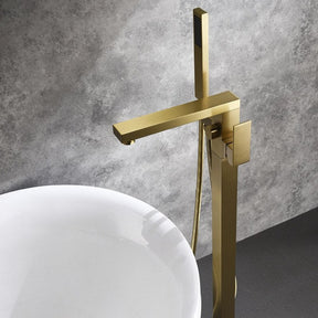 A modern brass Wasser™ Floor Mounted Bathtub Filler Shower Set by allfixture is installed against a gray wall. Water sprays horizontally from the handheld shower head attached to the fixture. The sleek, angular design and lever handle for controlling flow enhance its elegance. Below, the edge of a white bathtub is visible.