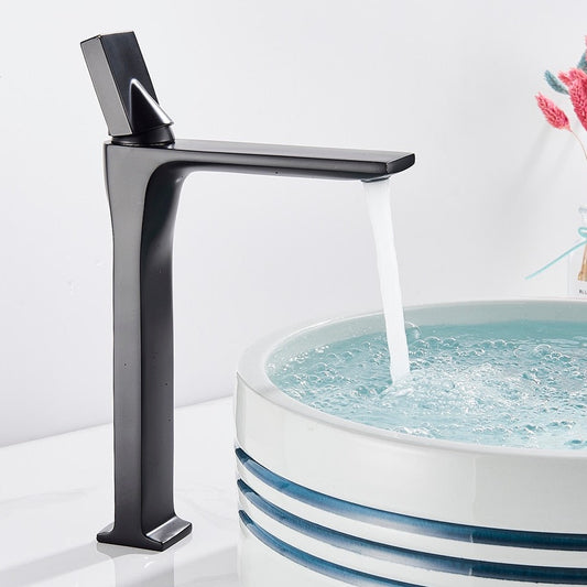 An AllFixture Deck Mounted Brass Single Handle Brass Basin Faucet in modern black matte is attached to a white counter. It is pouring water into a white, round basin with blue stripes near the rim. The counter is decorated with a vase of colorful dried flowers in the background.