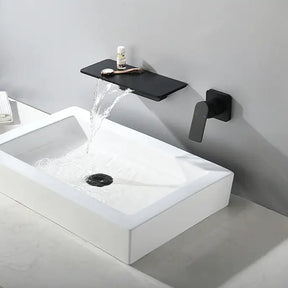 A modern bathroom sink with a minimalist design. The rectangular white sink has water pouring into it from the sleek, black Solid Brass Wall Mounted Waterfall Sink Faucet by AllFixture, which includes a stylish black handle. A shelf above the faucet holds a small container and a loofah. Towels are placed on the counter.