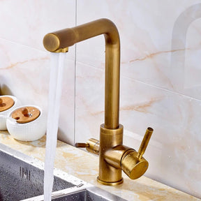 The Solid Brass Kitchen Faucet With Filtered Water Tap by allfixture graces the stainless steel sink with its sleek design, featuring a modern square-shaped neck and dual side handles for hot and cold water. Mounted on a wooden wall with a partially visible plant in the background, this faucet adds elegance to any culinary space.