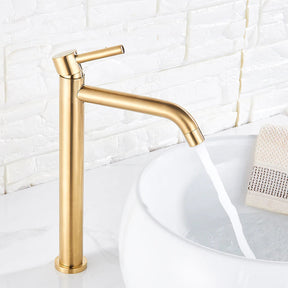 A sleek, modern Deck Mounted Bathroom Sink Faucet by Rozin in brushed gold stands next to a white, round basin sink on a white countertop. The background features a white textured tile wall. The faucet, much like an elegant shower head, has an elongated spout and a single lever handle.