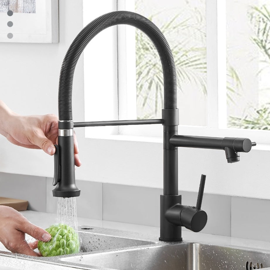 An AllFixture Single Handle Double Spout LED Kitchen Faucet with a sleek, modern black finish and a flexible spring neck dispenses water into a transparent glass. The faucet is mounted on a black countertop, set against wooden panels in the background. Natural light streams through a window framed in black in the background.