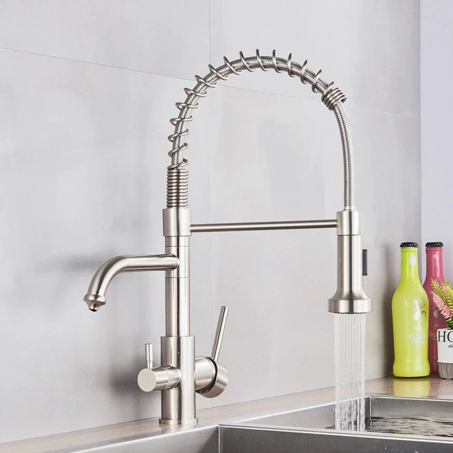 A MyQualife Double Handle Pull-Out Spout Kitchen Faucet with a high, curved design and flexible spring installation is installed over a stainless steel sink. The kitchen faucet features a versatile pull-out spray head and dual lever handles for water control. In the background, colorful bottles adorn the counter.