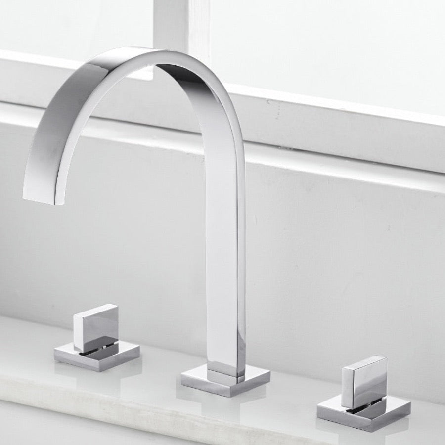 A minimalist black sink faucet from AllFixture, featuring an arching design, is mounted on a white countertop. The Double Handle Deck Mounted Faucet includes two lever handles—one on each side—with subtle red and blue indicators for hot and cold water respectively. In the background, a white window sill completes the scene.