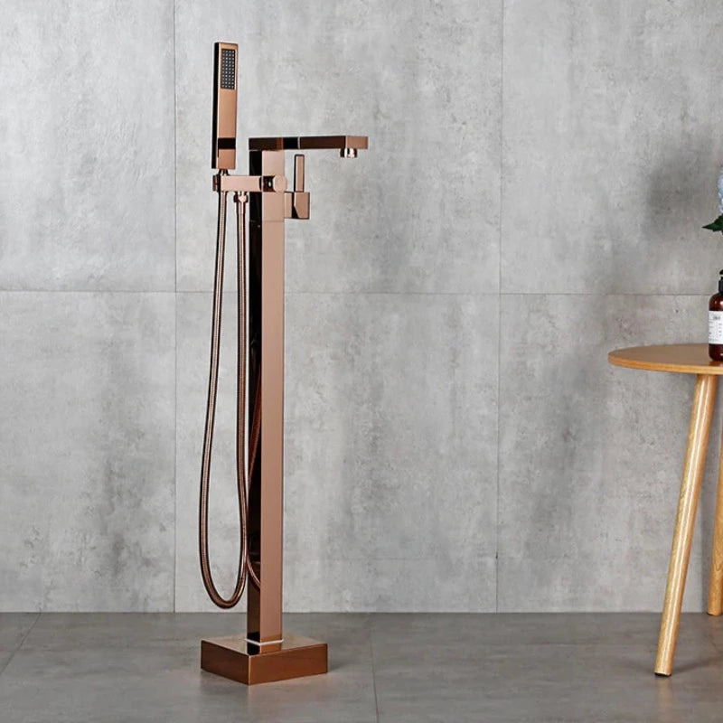 A sleek, freestanding, rose gold bathtub faucet from the Wasser™ collection by allfixture, set against a gray tile wall. The faucet showcases an angular design with a handheld shower attachment and hose, reminiscent of high-end bathroom fixtures. To the right, part of a wooden table with a bottle and some objects on it is visible.