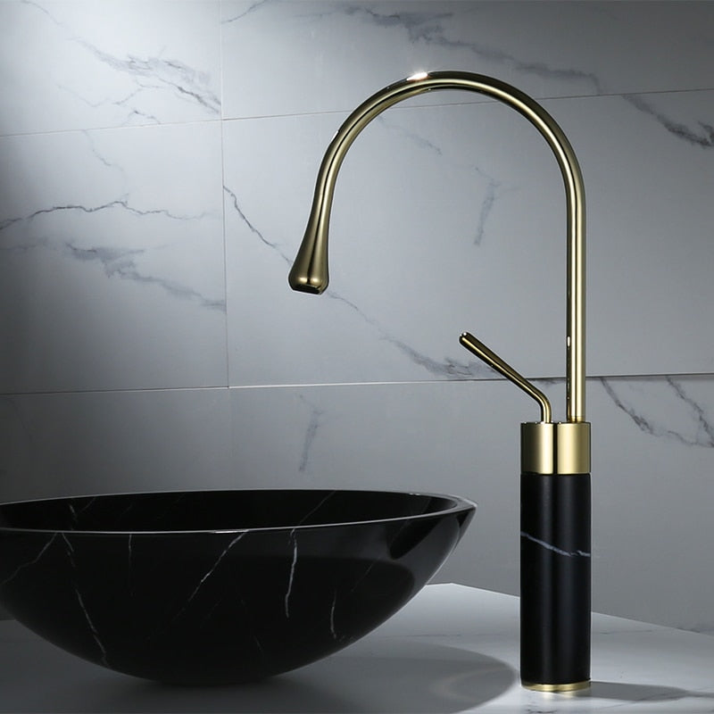 A sleek, modern bathroom sink setup featuring the Tiqui Solid Brass & Marble Bathroom Basin Faucet, showcasing a tall, curved gold design with a matching handle. Below the faucet is a stunning black marbled bowl sink. An elegant shower head complements the light grey tiles in the background, which have subtle marble veining.