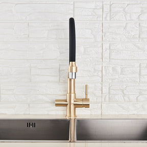 A modern kitchen sink with the sleek, gold-toned Solid Brass Kitchen Faucet With Filtered Water Tap by allfixture, featuring a black curved spout. The faucet is turned on, with water flowing over fresh produce in a stainless steel sink. In the background, there's a white brick wall and partially open window blinds.