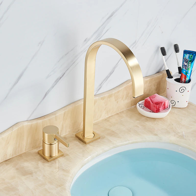 A brushed gold solid brass deck mounted bathroom sink faucet by allfixture showcases a modern high-arc spout and a single lever handle. Installed on a white countertop with a blurred plant and mirror in the background, this faucet creates a cohesive contemporary look, seamlessly complementing any kitchen sink faucet or shower head in matching finishes.