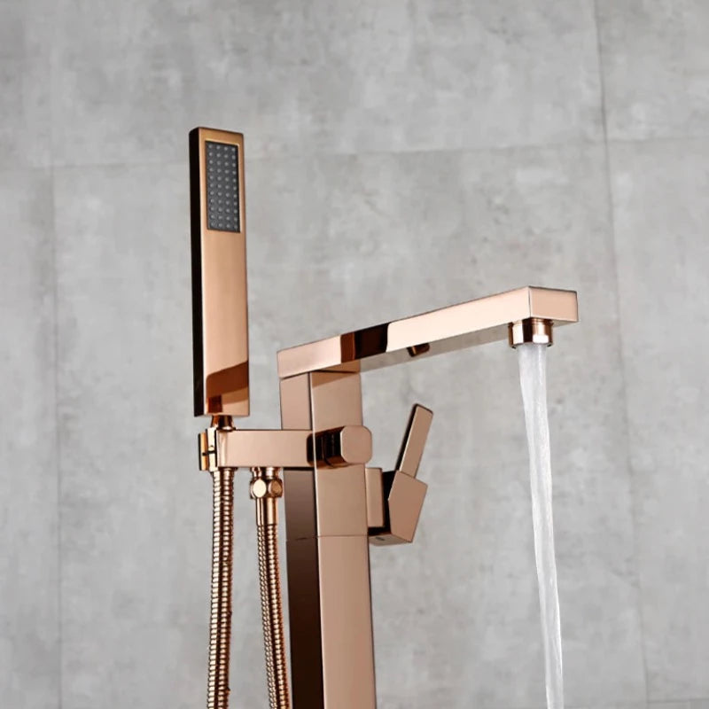 A sleek, freestanding, rose gold bathtub faucet from the Wasser™ collection by allfixture, set against a gray tile wall. The faucet showcases an angular design with a handheld shower attachment and hose, reminiscent of high-end bathroom fixtures. To the right, part of a wooden table with a bottle and some objects on it is visible.