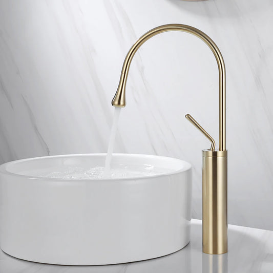 The Tiqui™ Single Handle Bathroom Sink Faucet in brushed gold, featuring a sleek curved high-arc spout, is elegantly mounted above a round white ceramic basin. Complementing the design is a matching shower head, set against a backdrop of gray marble-patterned tiles.