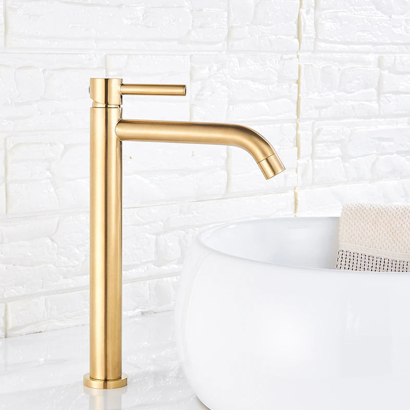 A sleek, modern Deck Mounted Bathroom Sink Faucet by Rozin in brushed gold stands next to a white, round basin sink on a white countertop. The background features a white textured tile wall. The faucet, much like an elegant shower head, has an elongated spout and a single lever handle.