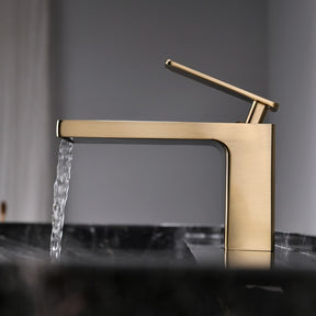 The Mike Jake Waterfall Style Solid Brass Bathroom Sink Faucet, with its modern rectangular design and angled lever handle, dispenses water into a white sink. In the background, a blurry geometric decoration and other bathroom essentials are visible. The faucet's minimalist and sleek aesthetic perfectly complements the setting.