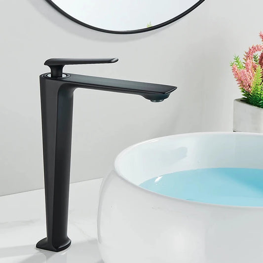 A Wasser™ Solid Brass Bathroom Sink Faucet by Rozin, featuring a sleek, angular design in modern black, dispenses water into a round, white vessel sink. In the background, a partial reflection is seen in a circular mirror, while a small plant adds a touch of greenery to the scene.