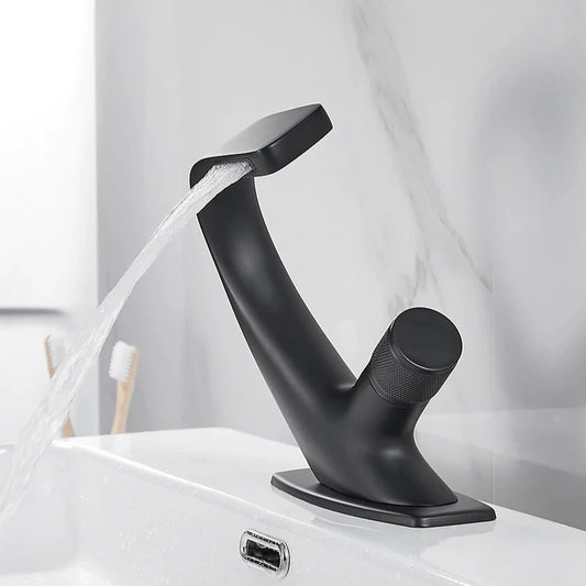 Close-up of an AllFixture Solid Brass Bathroom Basin Faucet with a modern, sleek black finish and angular design, installed on a white sink. Water is flowing from the faucet. In the background, a blurred image shows two toothbrushes in a holder on the countertop. The sink backdrop is a light-colored marble surface.