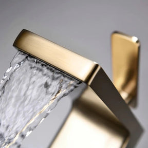 The Mike Jake Waterfall Style Solid Brass Bathroom Sink Faucet, with its modern rectangular design and angled lever handle, dispenses water into a white sink. In the background, a blurry geometric decoration and other bathroom essentials are visible. The faucet's minimalist and sleek aesthetic perfectly complements the setting.