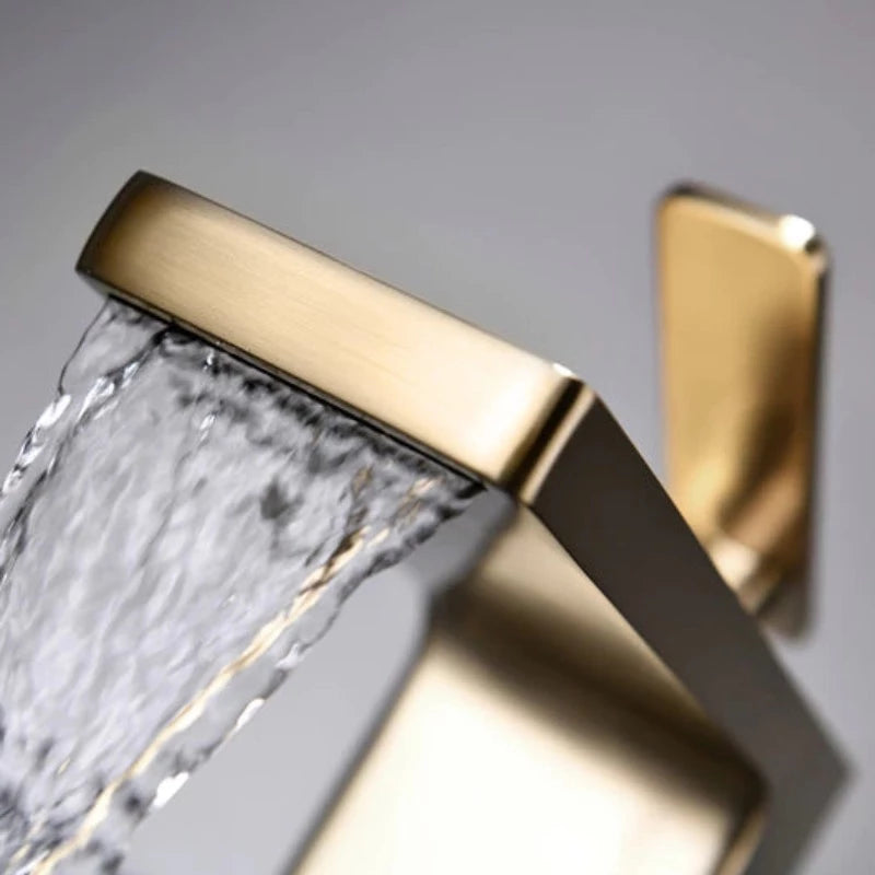 The Mike Jake Waterfall Style Solid Brass Bathroom Sink Faucet, with its modern rectangular design and angled lever handle, dispenses water into a white sink. In the background, a blurry geometric decoration and other bathroom essentials are visible. The faucet's minimalist and sleek aesthetic perfectly complements the setting.