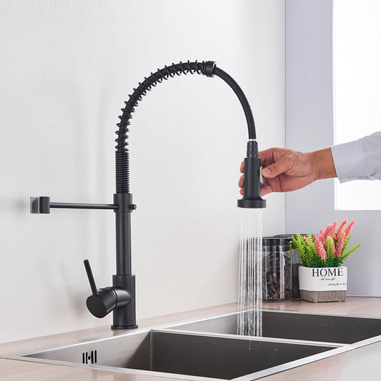 A person is holding the detachable spray head of the "Solid Brass Pull Out Spout Kitchen Sink Faucet" by allfixture, a modern black kitchen faucet featuring a curved, flexible spring design. Positioned over a double stainless steel sink, water streams from the spray head. Nearby, a plant and decorative "HOME" sign add charm to the scene.