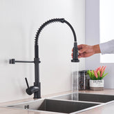 A person is holding the detachable spray head of the "Solid Brass Pull Out Spout Kitchen Sink Faucet" by allfixture, a modern black kitchen faucet featuring a curved, flexible spring design. Positioned over a double stainless steel sink, water streams from the spray head. Nearby, a plant and decorative "HOME" sign add charm to the scene.