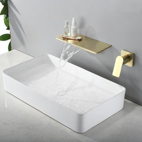 A minimalist bathroom sink features a rectangular white basin. Water flows gracefully from the Bagnox™ Wall Mounted Waterfall Brass Faucet by AllFixture, which doubles as a shelf holding two small bottles. The elegant setup is mounted on a light gray wall, near matching bathroom faucets, and part of a plant is visible on the left.