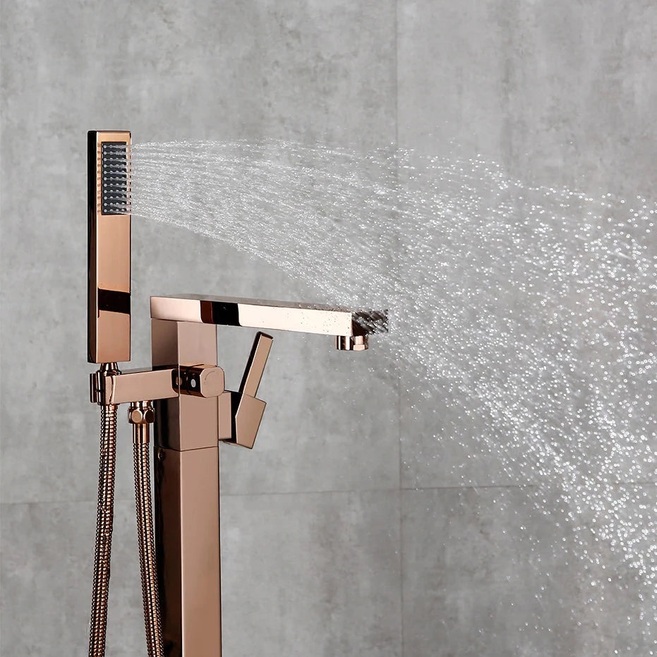 A sleek, freestanding, rose gold bathtub faucet from the Wasser™ collection by allfixture, set against a gray tile wall. The faucet showcases an angular design with a handheld shower attachment and hose, reminiscent of high-end bathroom fixtures. To the right, part of a wooden table with a bottle and some objects on it is visible.