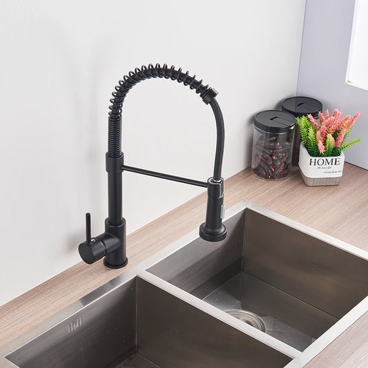 A person is holding the detachable spray head of the "Solid Brass Pull Out Spout Kitchen Sink Faucet" by allfixture, a modern black kitchen faucet featuring a curved, flexible spring design. Positioned over a double stainless steel sink, water streams from the spray head. Nearby, a plant and decorative "HOME" sign add charm to the scene.