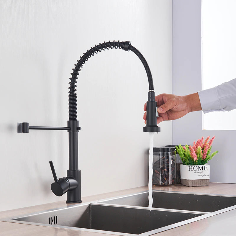 A person is holding the detachable spray head of the "Solid Brass Pull Out Spout Kitchen Sink Faucet" by allfixture, a modern black kitchen faucet featuring a curved, flexible spring design. Positioned over a double stainless steel sink, water streams from the spray head. Nearby, a plant and decorative "HOME" sign add charm to the scene.