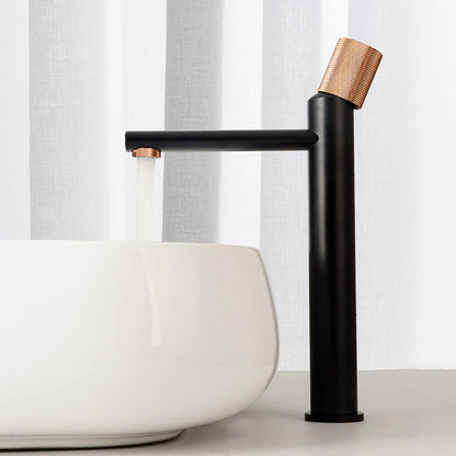 An AllFixture Wasser™ deck-mounted bathroom faucet, featuring a sleek black finish and a gold-toned handle, is elegantly mounted over a modern, minimalist white vessel sink. Water flows seamlessly from the faucet against a backdrop of either a white curtain or wall, enhancing the clean and contemporary aesthetic.