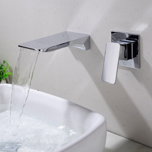 A modern, wall-mounted bathroom sink faucet by AllFixture, made of solid brass and featuring a flat spout, is dispensing water into a white rectangular sink. The chrome faucet handle is positioned to the right of the spout. A small green plant is partially visible to the left of the sink.