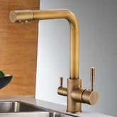 The Solid Brass Kitchen Faucet With Filtered Water Tap by allfixture graces the stainless steel sink with its sleek design, featuring a modern square-shaped neck and dual side handles for hot and cold water. Mounted on a wooden wall with a partially visible plant in the background, this faucet adds elegance to any culinary space.