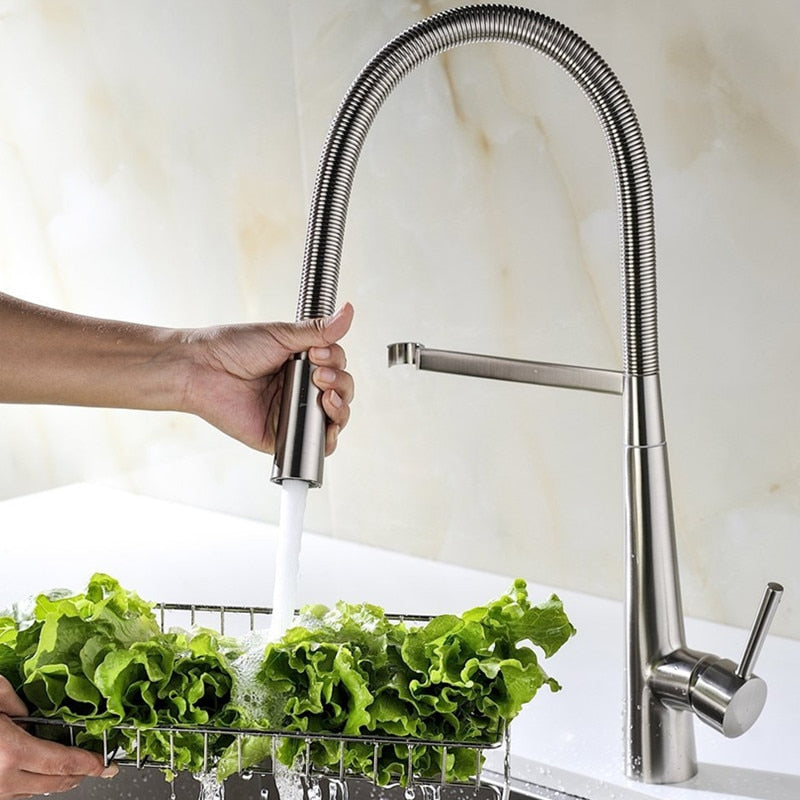 A person rinses fresh green lettuce under an allfixture Pull Down 2 Function Sprayer Brass Kitchen Sink Faucet with a tall, flexible design and pull-down spray head. The stainless steel faucet features a single lever handle, and the water flows into a white sink beneath.