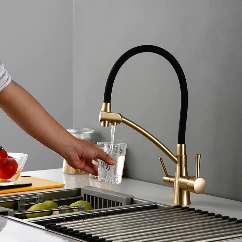 A person is using the Wasser™ Solid Brass Kitchen Faucet With Filtered Water Tap by allfixture to wash green apples in a sink. The countertop features a cutting board with a tomato, and jars of pasta are visible in the background. The overall kitchen decor is minimalist and contemporary, highlighting the modern design of the sleek gold and black kitchen faucet.