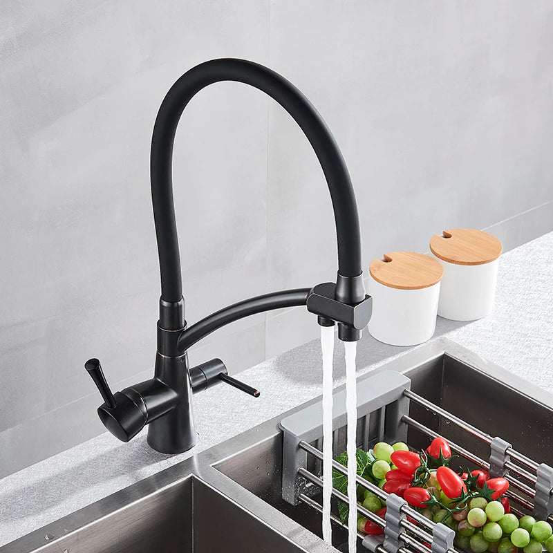 A person holds the spout of the Rozin Deck Mounted Brass Kitchen Faucet in black, directing a stream of water into a stainless steel sink filled with vegetables. The background features a sleek, minimalist kitchen with two white containers on the countertop.