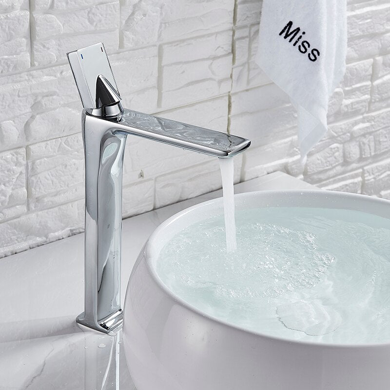 An AllFixture Deck Mounted Brass Single Handle Brass Basin Faucet in modern black matte is attached to a white counter. It is pouring water into a white, round basin with blue stripes near the rim. The counter is decorated with a vase of colorful dried flowers in the background.