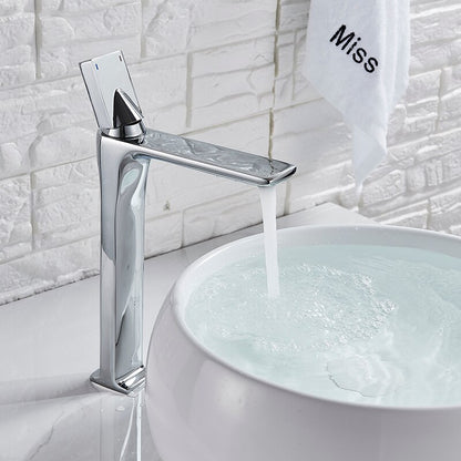 An AllFixture Deck Mounted Brass Single Handle Brass Basin Faucet in modern black matte is attached to a white counter. It is pouring water into a white, round basin with blue stripes near the rim. The counter is decorated with a vase of colorful dried flowers in the background.