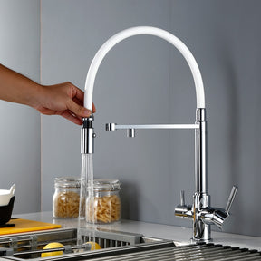 A person's hand is adjusting the spray nozzle of the Bagnolux 3-Function Pull Down Spout Kitchen Sink Faucet with a flexible, white arched hose. The faucet, featuring a ceramic valve and chrome finish, is installed above a stainless steel sink. In the background, there are pasta jars and a dish drying rack.