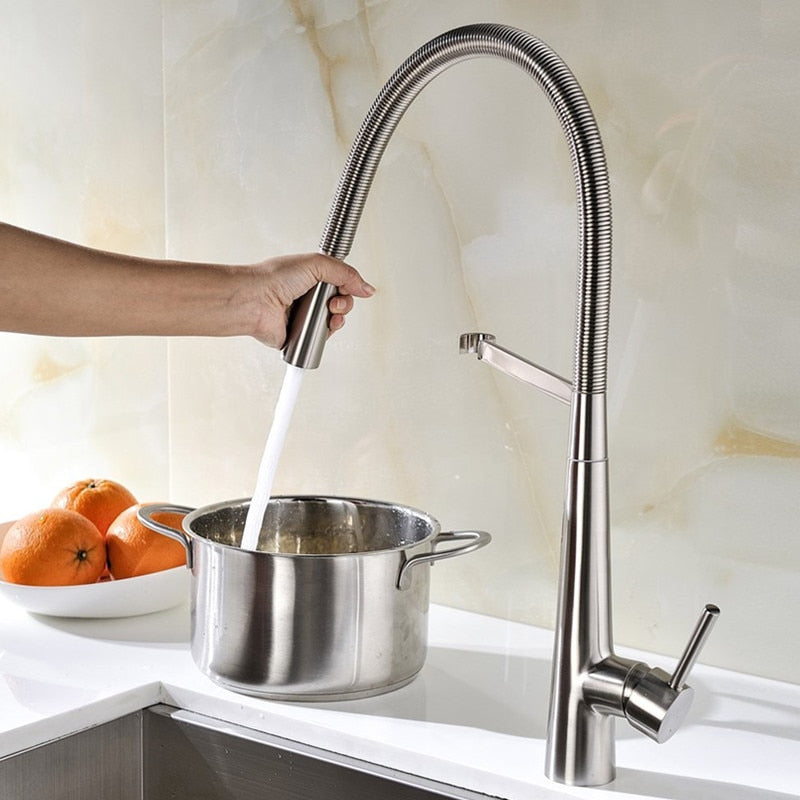 A person rinses fresh green lettuce under an allfixture Pull Down 2 Function Sprayer Brass Kitchen Sink Faucet with a tall, flexible design and pull-down spray head. The stainless steel faucet features a single lever handle, and the water flows into a white sink beneath.