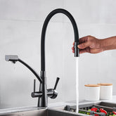 A person holds the spout of the Rozin Deck Mounted Brass Kitchen Faucet in black, directing a stream of water into a stainless steel sink filled with vegetables. The background features a sleek, minimalist kitchen with two white containers on the countertop.