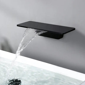 A minimalist bathroom sink features a rectangular white basin. Water flows gracefully from the Bagnox™ Wall Mounted Waterfall Brass Faucet by AllFixture, which doubles as a shelf holding two small bottles. The elegant setup is mounted on a light gray wall, near matching bathroom faucets, and part of a plant is visible on the left.