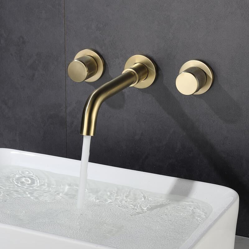 The Tiqui Solid Brass Wall Mounted Bathroom Faucet in brushed gold, which features two round handles and a stylish spout, pours water into a white rectangular sink filled with water against a dark gray tiled wall.