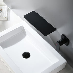 A modern bathroom sink setup featuring the AllFixture Solid Brass Wall Mounted Waterfall Sink Faucet extending horizontally from the wall in a gold finish. Water flows gracefully from the faucet into a rectangular white basin. Two small bottles are placed on the faucet shelf above the cascading water. The wall and countertop are decorated in muted tones.