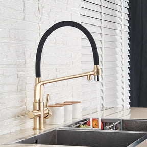 A modern kitchen sink with the sleek, gold-toned Solid Brass Kitchen Faucet With Filtered Water Tap by allfixture, featuring a black curved spout. The faucet is turned on, with water flowing over fresh produce in a stainless steel sink. In the background, there's a white brick wall and partially open window blinds.