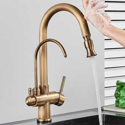 A person uses the Rozin Touch Sensor Antique Brass Kitchen Faucet with Filtered Water Tap to wash their hands. The elegant faucet features dual handles and a high-arc spout, providing a modern touch to the kitchen sink area, which is immaculate against a backdrop of white and green-striped walls.