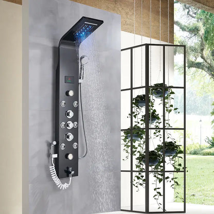 A stylish Morsale.com 6-Function Wall Shower Panel With Massage Jets in Charcoal is installed on a tiled wall. Water is flowing from the handheld shower faucet. To the right, a tall glass and metal plant stand holds several potted plants, with greenery visible outside.