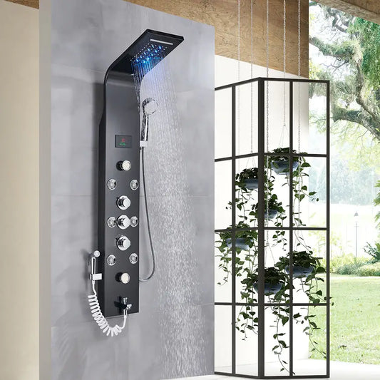 A stylish Morsale.com 6-Function Wall Shower Panel With Massage Jets in Charcoal is installed on a tiled wall. Water is flowing from the handheld shower faucet. To the right, a tall glass and metal plant stand holds several potted plants, with greenery visible outside.