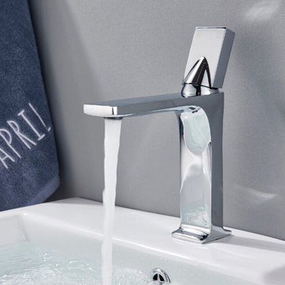 An AllFixture Deck Mounted Brass Single Handle Brass Basin Faucet in modern black matte is attached to a white counter. It is pouring water into a white, round basin with blue stripes near the rim. The counter is decorated with a vase of colorful dried flowers in the background.