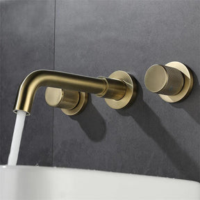 The Tiqui Solid Brass Wall Mounted Bathroom Faucet in brushed gold, which features two round handles and a stylish spout, pours water into a white rectangular sink filled with water against a dark gray tiled wall.