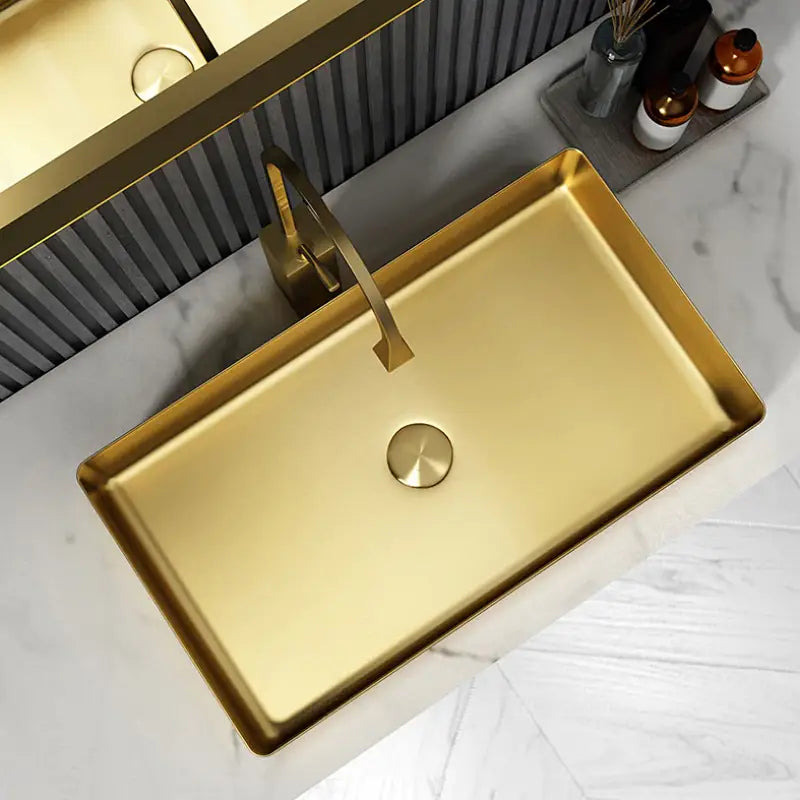 A modern bathroom showcasing the SUS304 Stainless Steel Rectangle Bathroom Sink in Brushed Gold by Mike Jake, set on a white marble countertop. Above the sink is a matching gold rectangular faucet. The wall behind features vertical gray stripes and a large rectangular mirror. To the left of the sink, two brown bottles and a reed diffuser complement other sleek bathroom faucets.