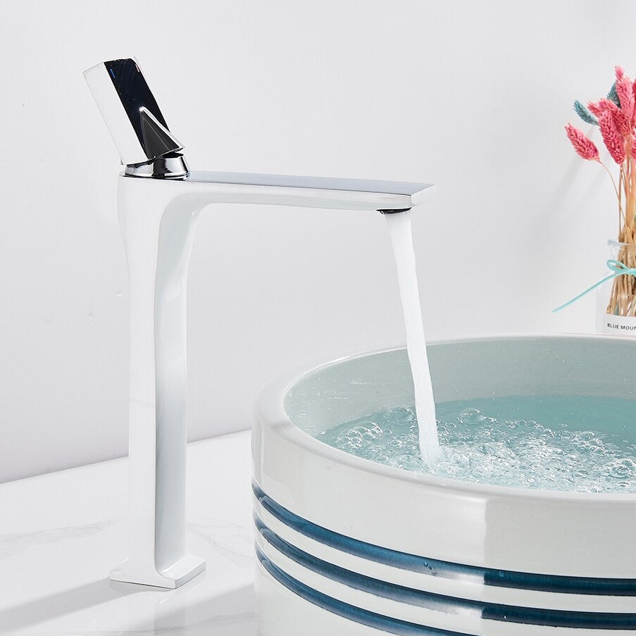 An AllFixture Deck Mounted Brass Single Handle Brass Basin Faucet in modern black matte is attached to a white counter. It is pouring water into a white, round basin with blue stripes near the rim. The counter is decorated with a vase of colorful dried flowers in the background.