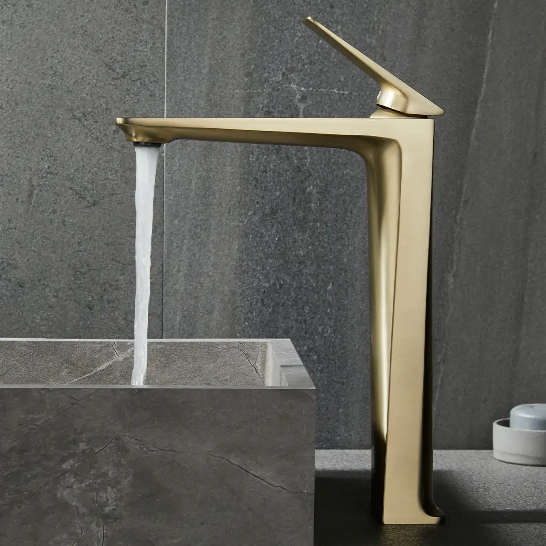 A modern, gold-colored Bagnolux Single Handle Brass Bathroom Sink Faucet with a sleek design is mounted on a grey stone countertop, pouring water into a matching rectangular grey stone sink. The backdrop features a textured grey wall, completing this elegant and cohesive look.