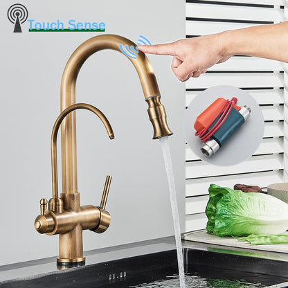 A person uses the Rozin Touch Sensor Antique Brass Kitchen Faucet with Filtered Water Tap to wash their hands. The elegant faucet features dual handles and a high-arc spout, providing a modern touch to the kitchen sink area, which is immaculate against a backdrop of white and green-striped walls.